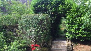 Una escalera de piedra que sube a un jardín con abundante vegetación y un jarrón de flores rojas.