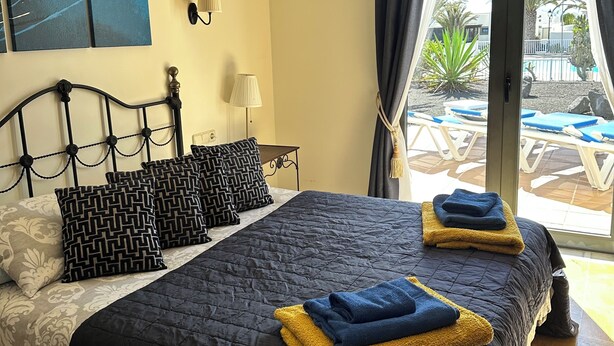 Een slaapkamer met een bed, kussens, een nachtkastje, een lamp en uitzicht op een buitenzwembad.