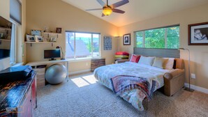 Ein Schlafzimmer mit einem Bett, einem Schreibtisch, einem Computer, einem Deckenventilator und einem Fenster mit Blick auf Grünflächen.