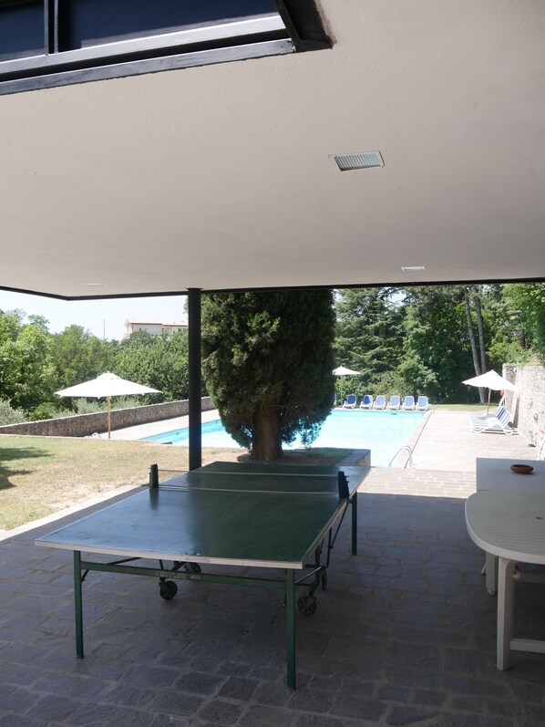 An outdoor table tennis table under a covered area with a pool and lounge chairs in the background.