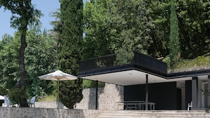 Ein moderner Poolbereich im Freien mit einer Steinmauer, einer überdachten Sitzecke und Blick auf hohe Bäume.