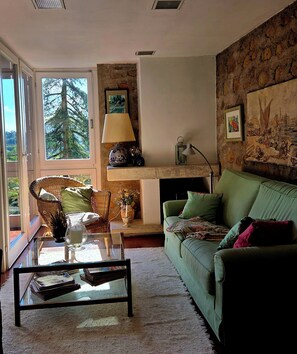 Ein Wohnzimmer mit einem grünen Sofa, einem Korbstuhl, einem Glastisch, einem Kamin und einem großen Fenster mit Blick auf Bäume.