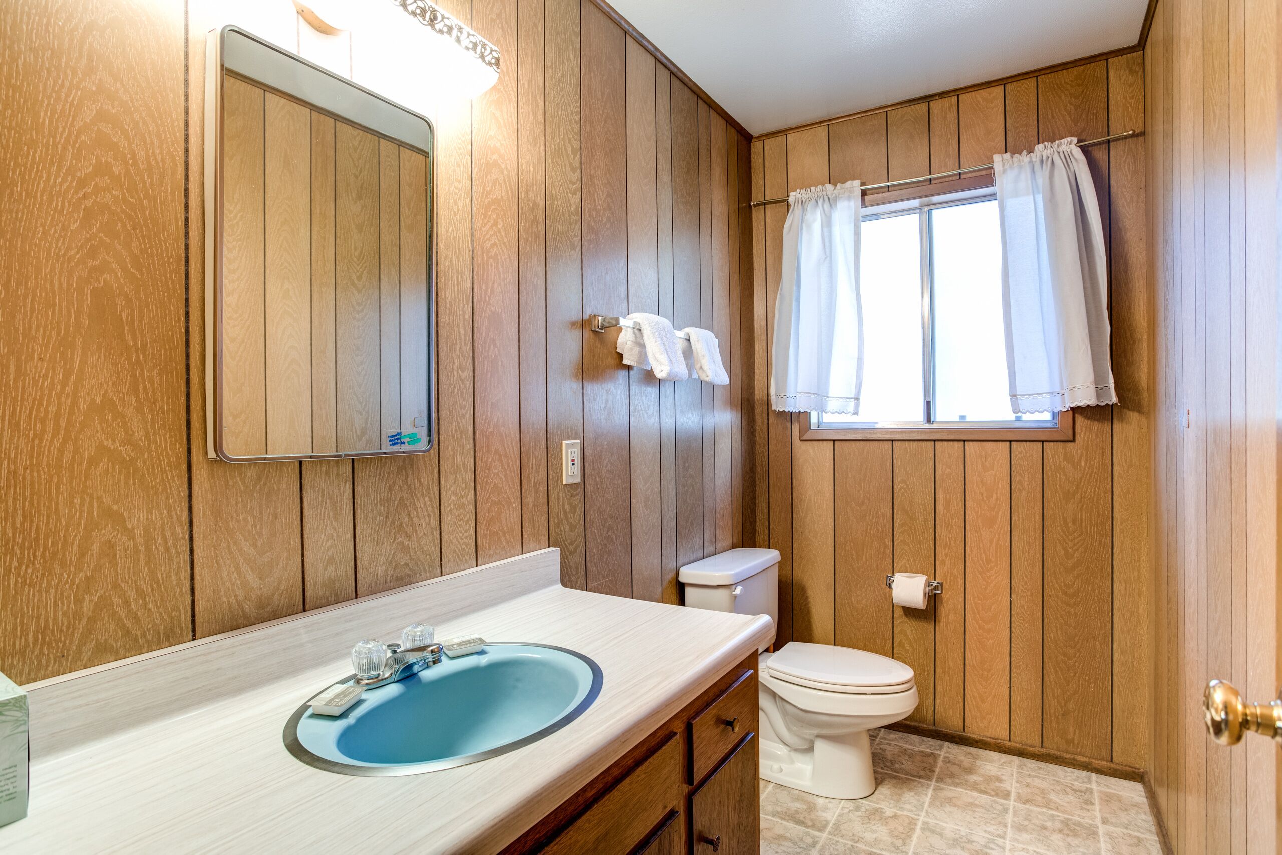A bathroom with wooden paneling, a sink with a mirror, a toilet, and a window with curtains.