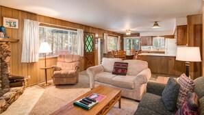 A cozy living room with a fireplace, a sofa, a coffee table, and a dining area with a kitchen in the background.