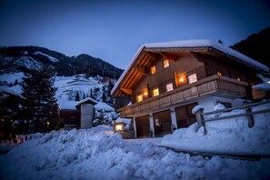 A wooden chalet with multiple windows and a balcony, surrounded by snow-covered hills and trees.