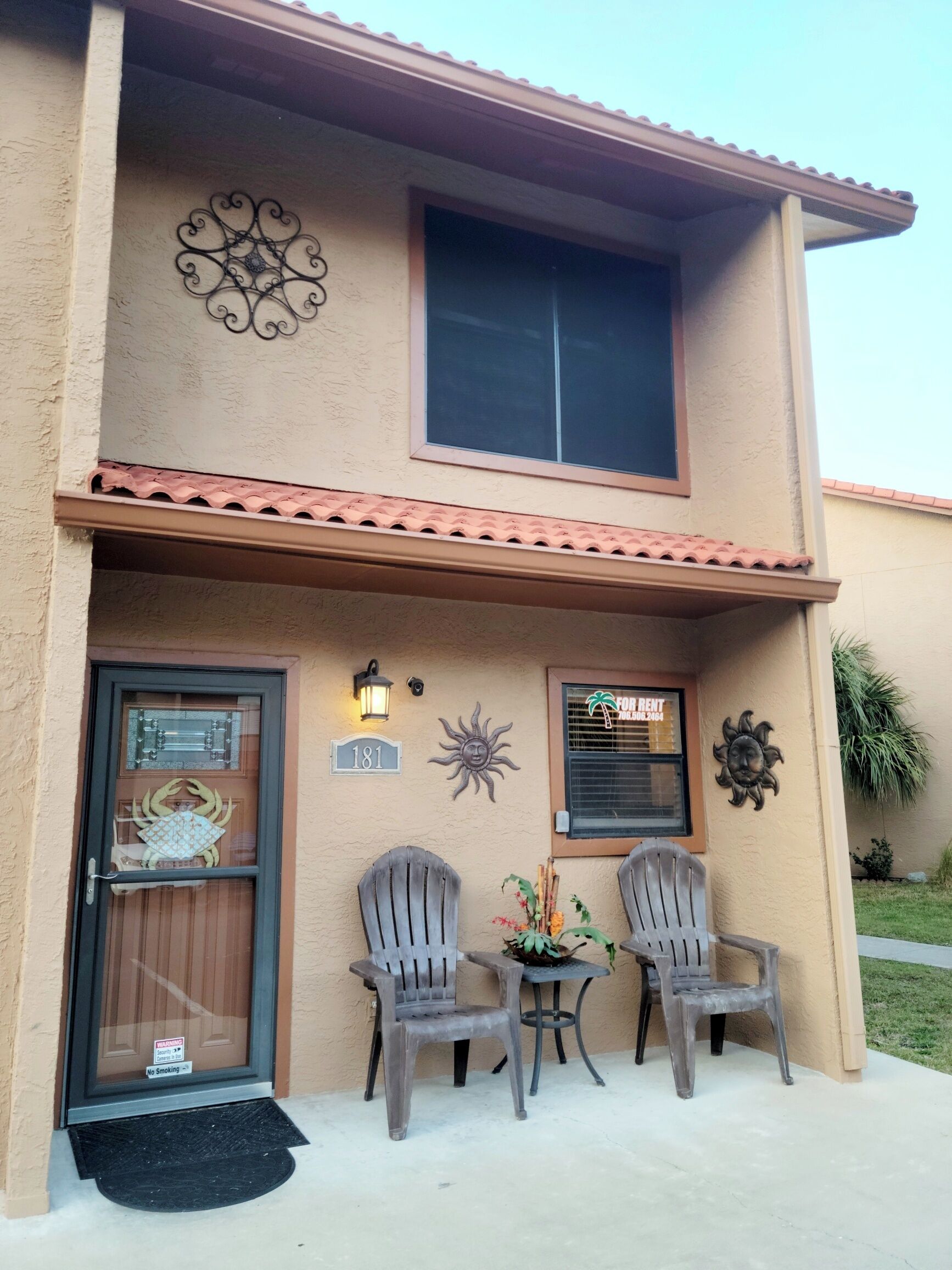 A two-story building with a beige exterior, a large window on the upper floor, and a door with a decorative crab design. There are two chairs outside and a small table with a plant.