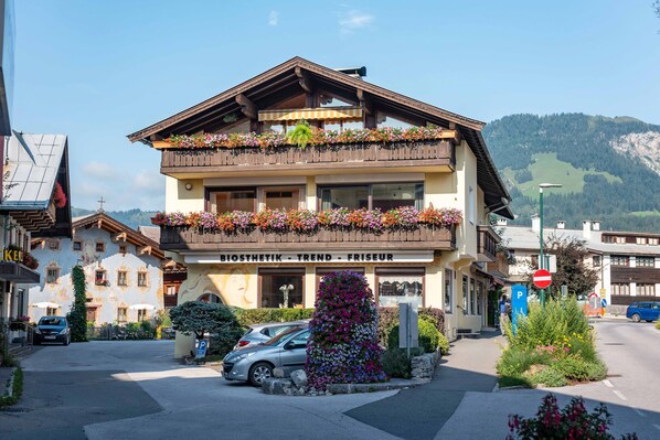 A two-story building with balconies adorned with flowers, a sign reading "BIOSYNTEKTIK - TREND - FRISEUR," and a parking area with cars.