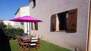 Um pátio com mesa e cadeiras de madeira, um guarda-sol roxo e um pequeno gramado.