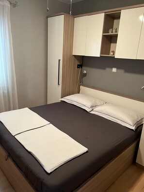 A single bed with a dark mattress and white linens, a wooden wardrobe with a mirror, and a small shelf with decorative items.
