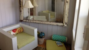 A room with a white wicker sofa with green cushions, a small ottoman with a book, a wicker basket, and a mirror with a lamp.