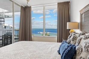 Habitación de hotel con un gran ventanal que ofrece vistas al mar y un muelle. Se observa una cama con cubierta blanca, una lámpara y una silla.