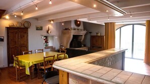 Uma sala de jantar com uma mesa comprida, cadeiras, lareira e uma área de cozinha com janela.