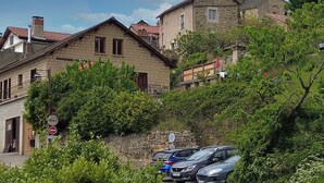 Um edifício histórico de pedra com uma torre de sino, rodeado por vegetação exuberante e carros estacionados.