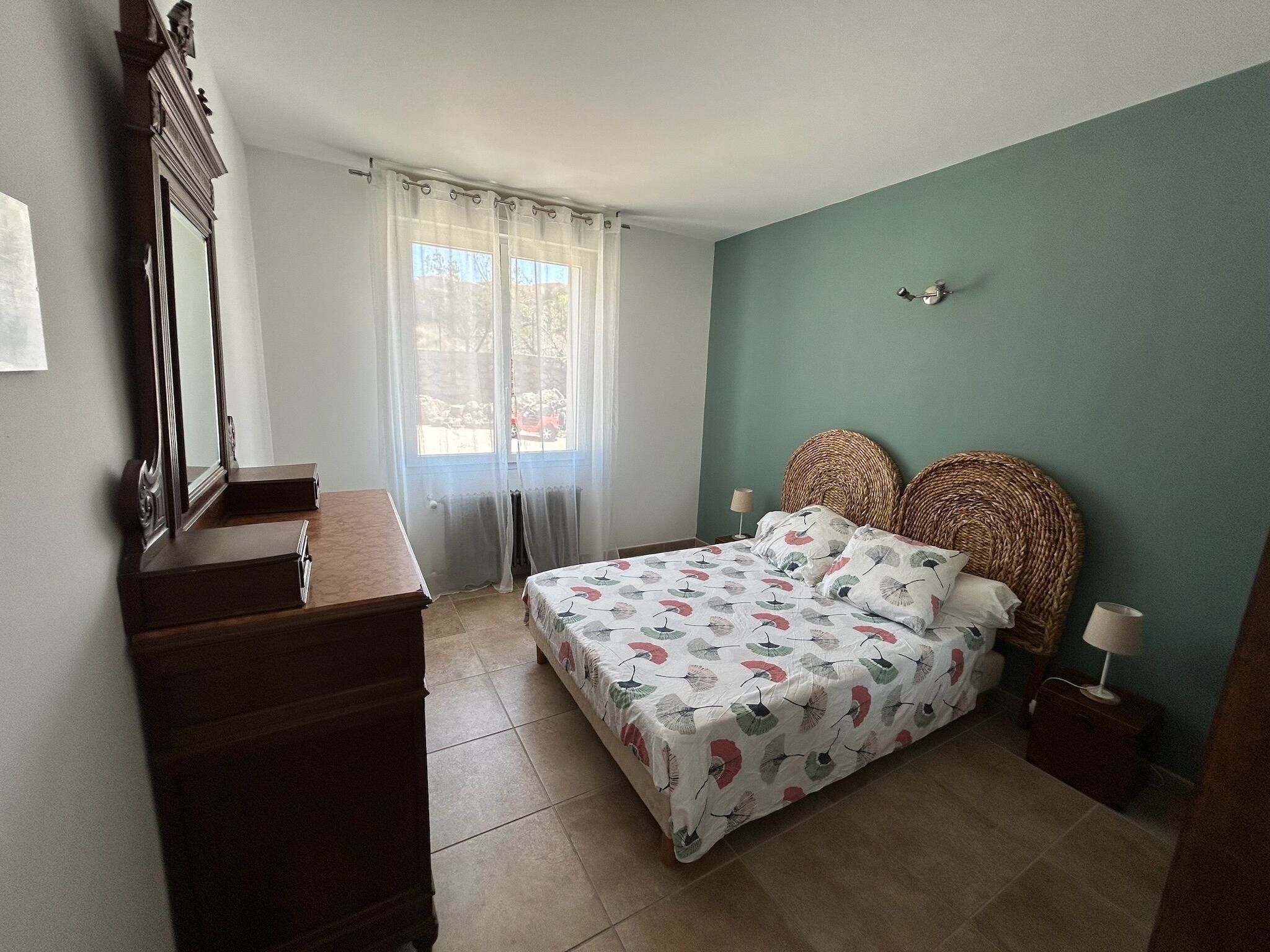 Un dormitorio con cama, cómoda, espejo, ventana con cortinas y una lámpara de pared.