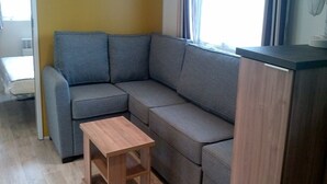 A corner room with a grey sofa, a wooden side table, and a wooden cabinet. - 3 Bedroom mobile home Siblu Domaine de Kerlann  Brittany France 131 (Pont-Aven)