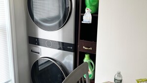 A compact laundry room with two stacked front-loading washing machines, a small table with a striped tablecloth, and a chair. - One Bedroom Apartment with Separate Bunk Bed Area (Shrewsbury)