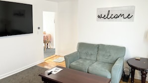 A living room with a green sofa, a TV, a welcome sign, and a small table. - One Bedroom Apartment with Separate Bunk Bed Area (Shrewsbury)