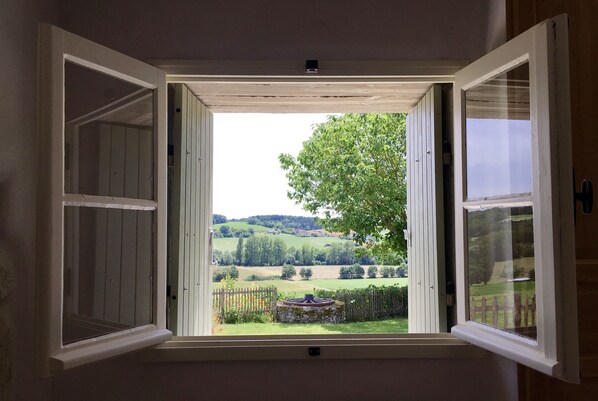 Una ventana abierta con vistas a un paisaje verde, un árbol y una construcción de piedra.