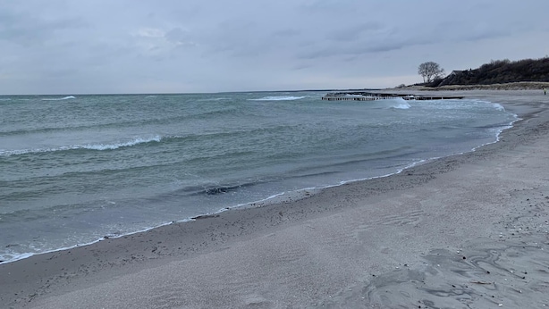Une plage avec une jetée en bois qui s’avance dans l’eau, un rivage sablonneux et un ciel nuageux.