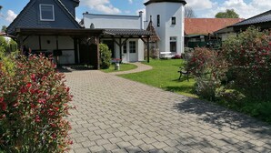 Une maison avec une allée pavée menant à un porche couvert, une petite structure ressemblant à une tour et un jardin agrémenté de plantes à fleurs.
