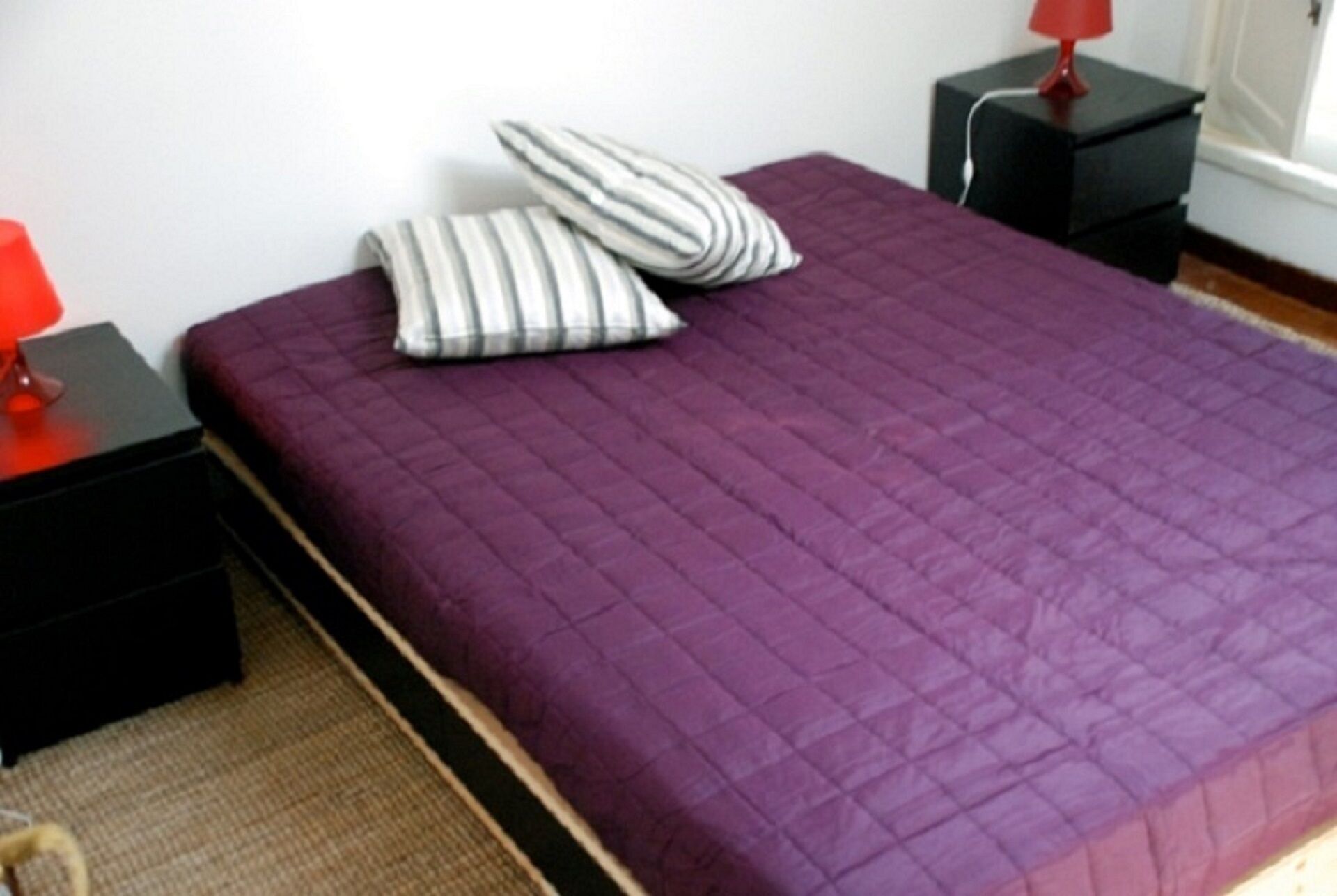Ein Bett mit einer violetten Decke, zwei gestreiften Kissen und einer roten Lampe auf einem schwarzen Nachttisch.