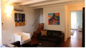 A living room with a white armchair, a dark sofa, and two colorful abstract paintings on the walls. - Fully furnished apartment, with all comforts. ALL INCLUSIVE (Siracusa)
