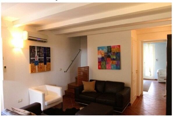 A living room with a white armchair, a dark sofa, and two colorful abstract paintings on the walls.
