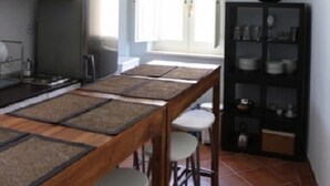 A kitchen with a long wooden table, bar stools, a black cabinet with dishes, and a window with blinds. - Fully furnished apartment, with all comforts. ALL INCLUSIVE (Siracusa)