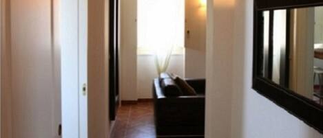 A narrow hallway with a tiled floor, a sofa in the background, and a table with various items on it.