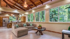 A spacious living area with a wooden ceiling, large windows with a view of greenery, a kitchen in the background, and a cozy seating arrangement. - Casa Bamboo | 4-Bedroom Colonial Jungle Villa | Private Pool | Manuel Antonio (Manuel Antonio)