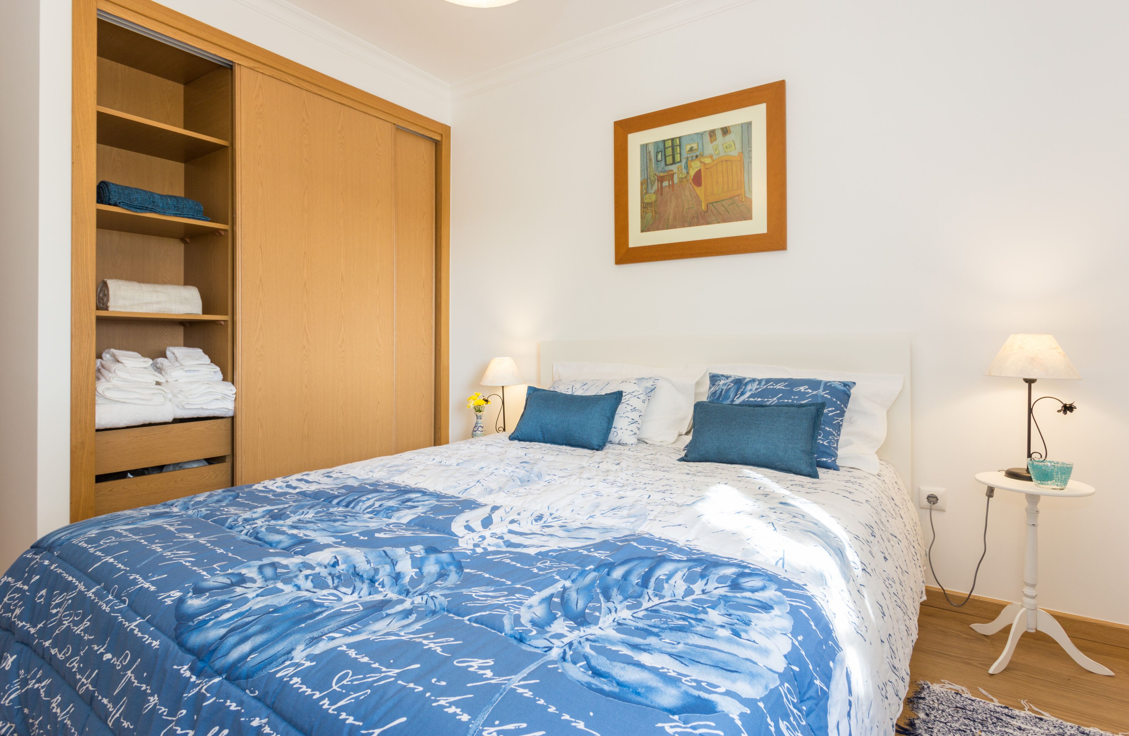 A bedroom with a large bed, a wooden wardrobe, a framed picture on the wall, and a bedside table with a lamp.