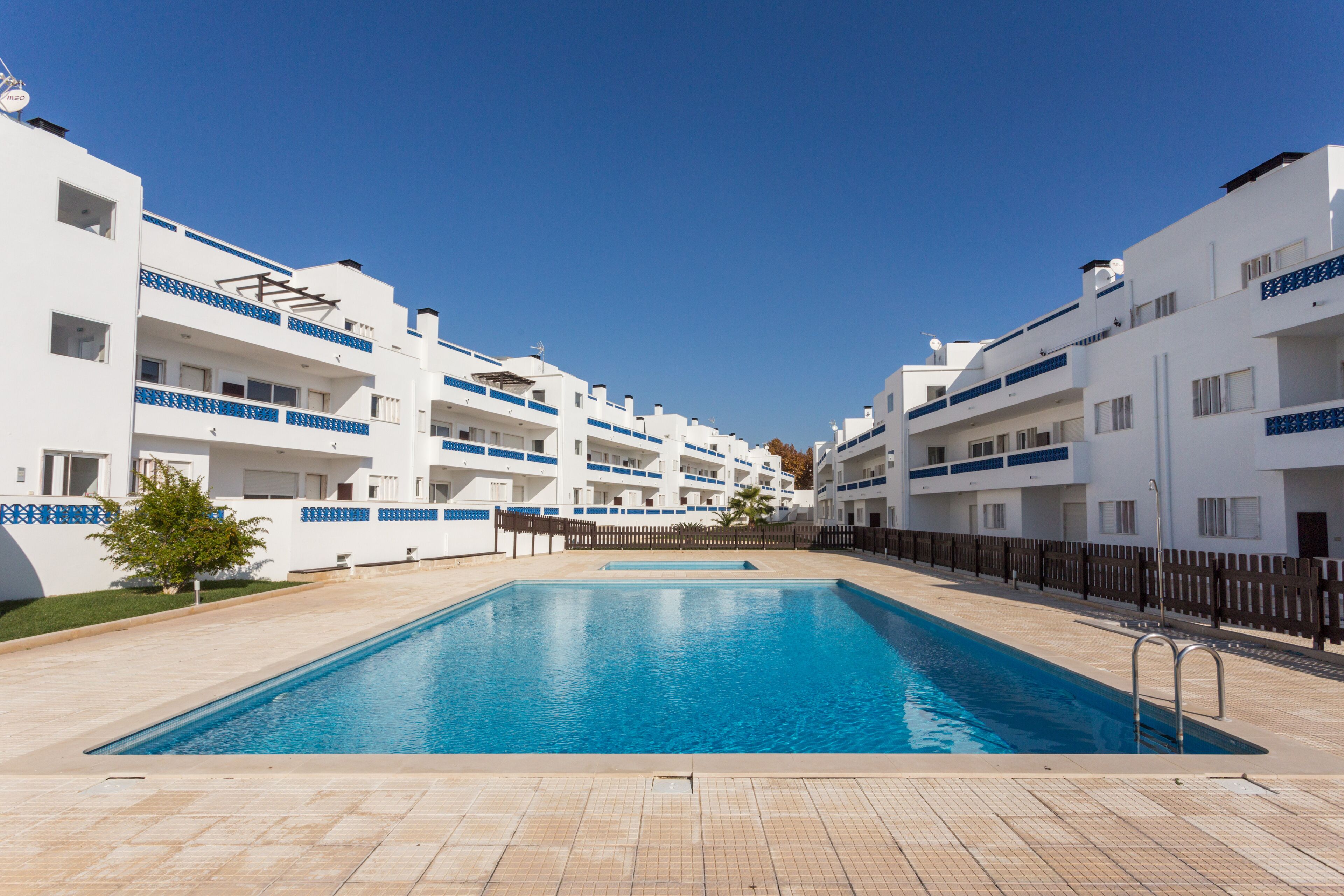 A swimming pool surrounded by white buildings with blue accents, a clear blue sky, and a tiled deck.