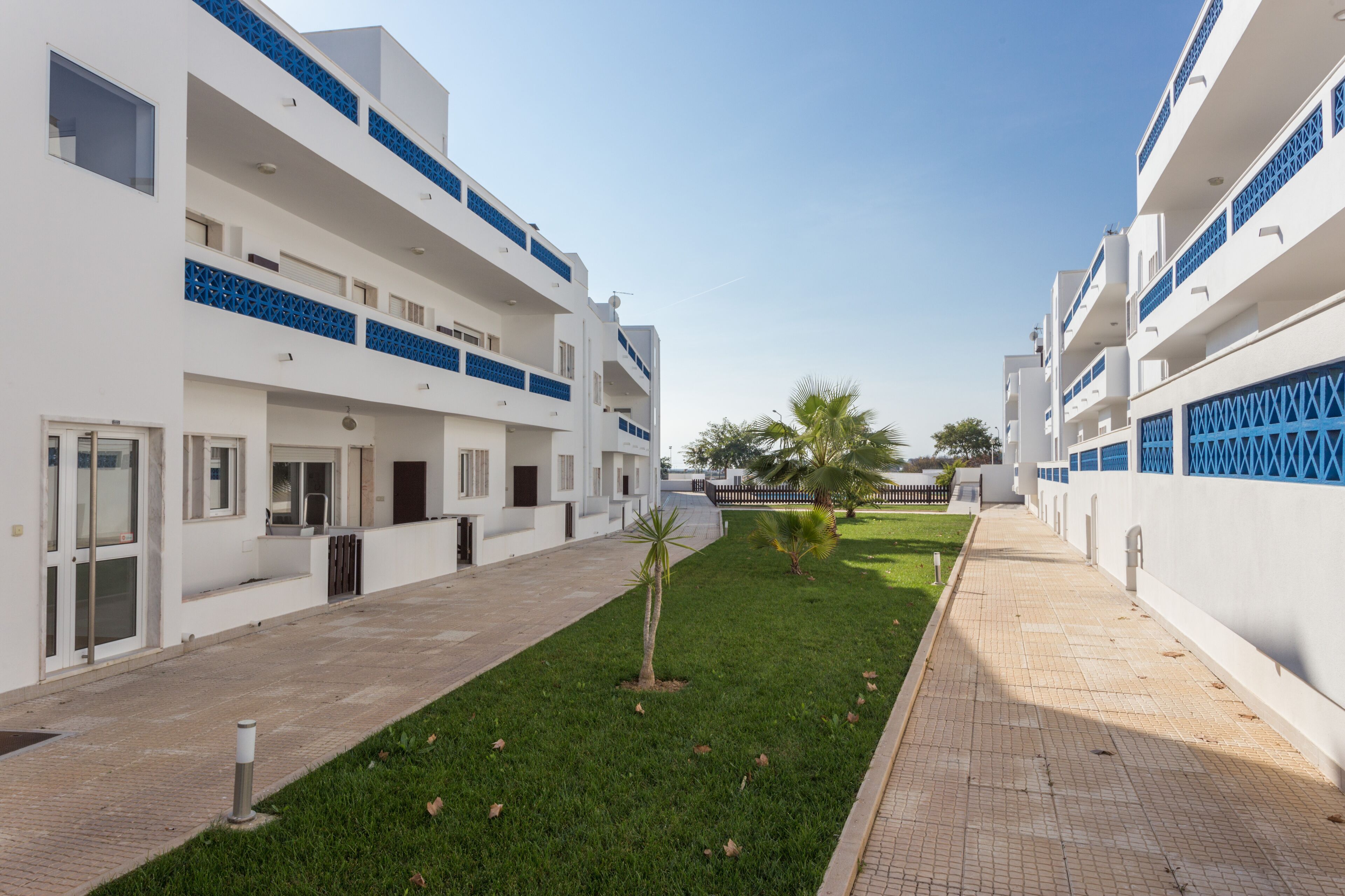 Un bâtiment moderne aux façades blanches et bleues, doté d’une cour intérieure avec pelouse et palmiers, ainsi que d’un chemin pavé.