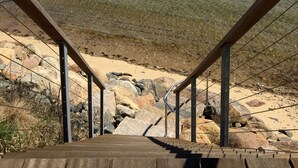 Uma escadaria de madeira que desce até uma praia rochosa, com águas cristalinas e um litoral distante.