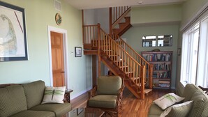Uma sala de estar ampla com escada de madeira, sofás com padrão verde, uma mesa de centro de vidro e uma estante cheia de livros.