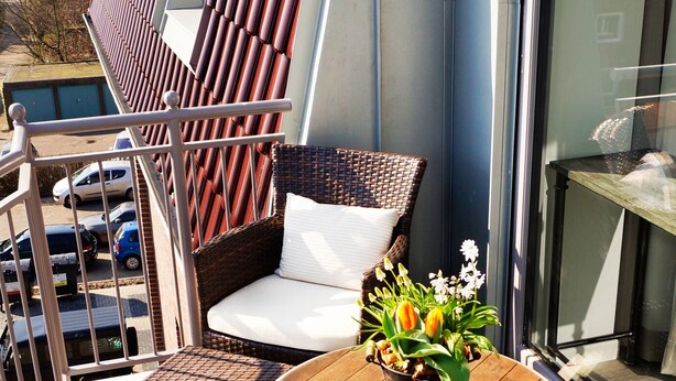 A balcony with wicker furniture, a round table with a potted plant, and a view of the city through large windows.