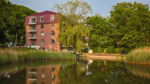 A multi-story brick building with a red roof, reflecting in a calm body of water.