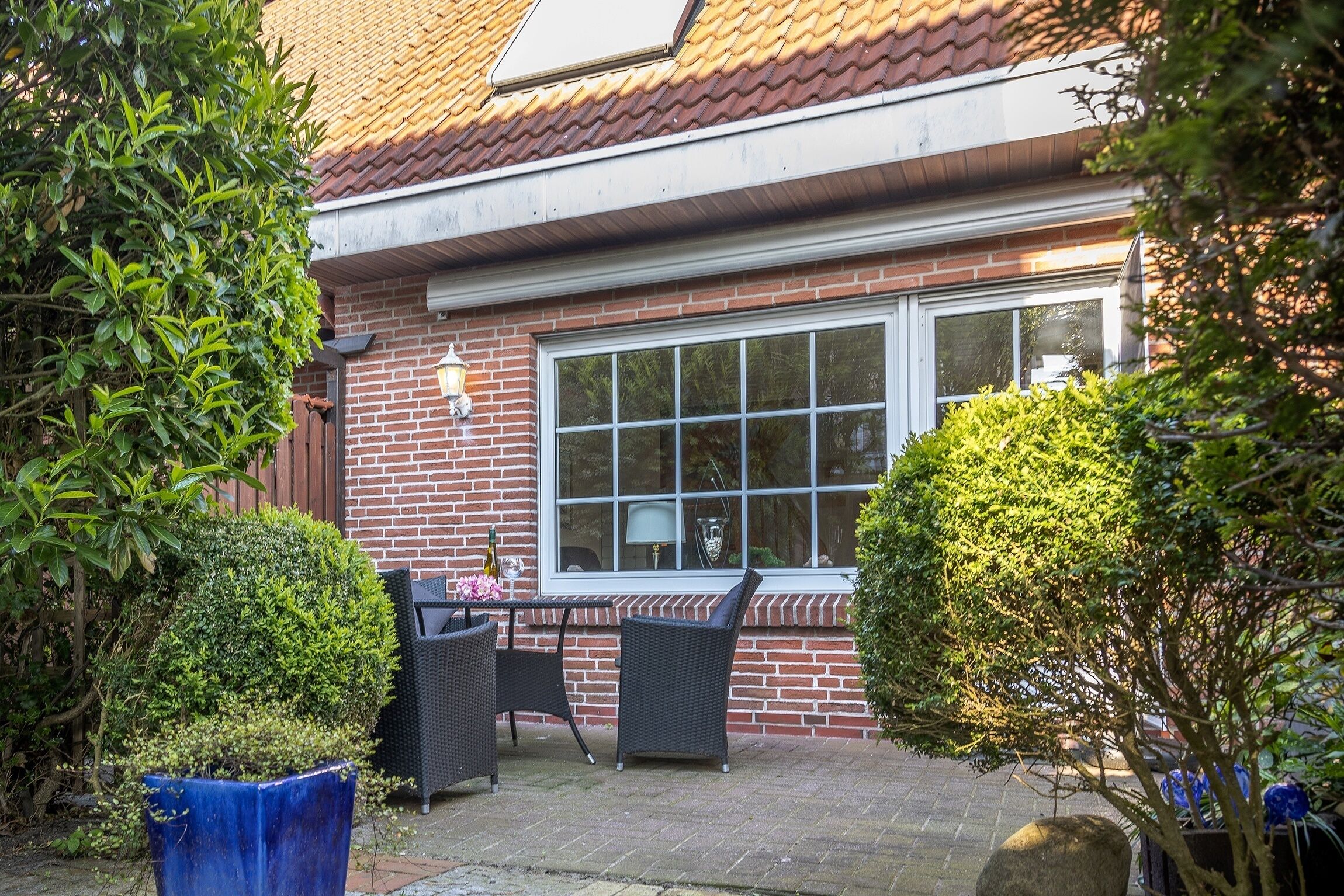 Une maison en briques avec un toit en tuiles, une grande fenêtre et un patio aménagé avec des meubles en osier et des plantes en pot.
