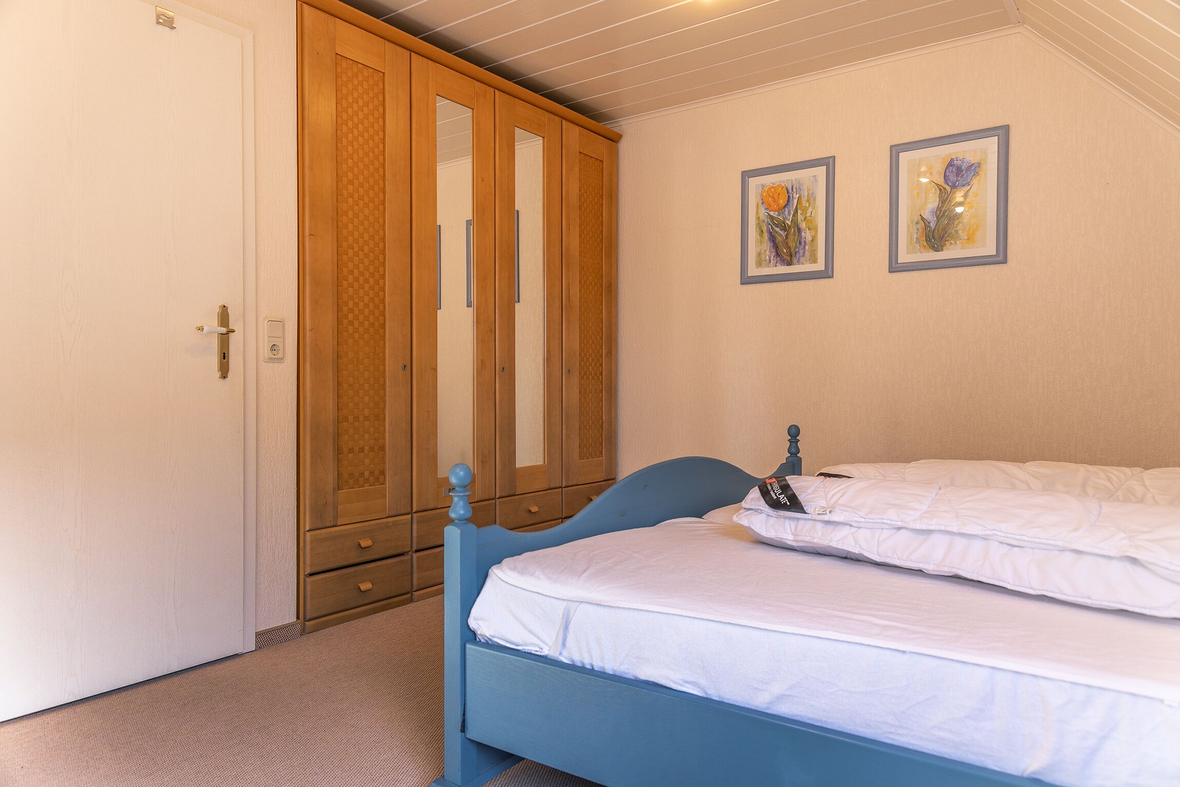 A bedroom with a blue bed, white bedding, a wooden wardrobe, and two framed artworks on the wall.