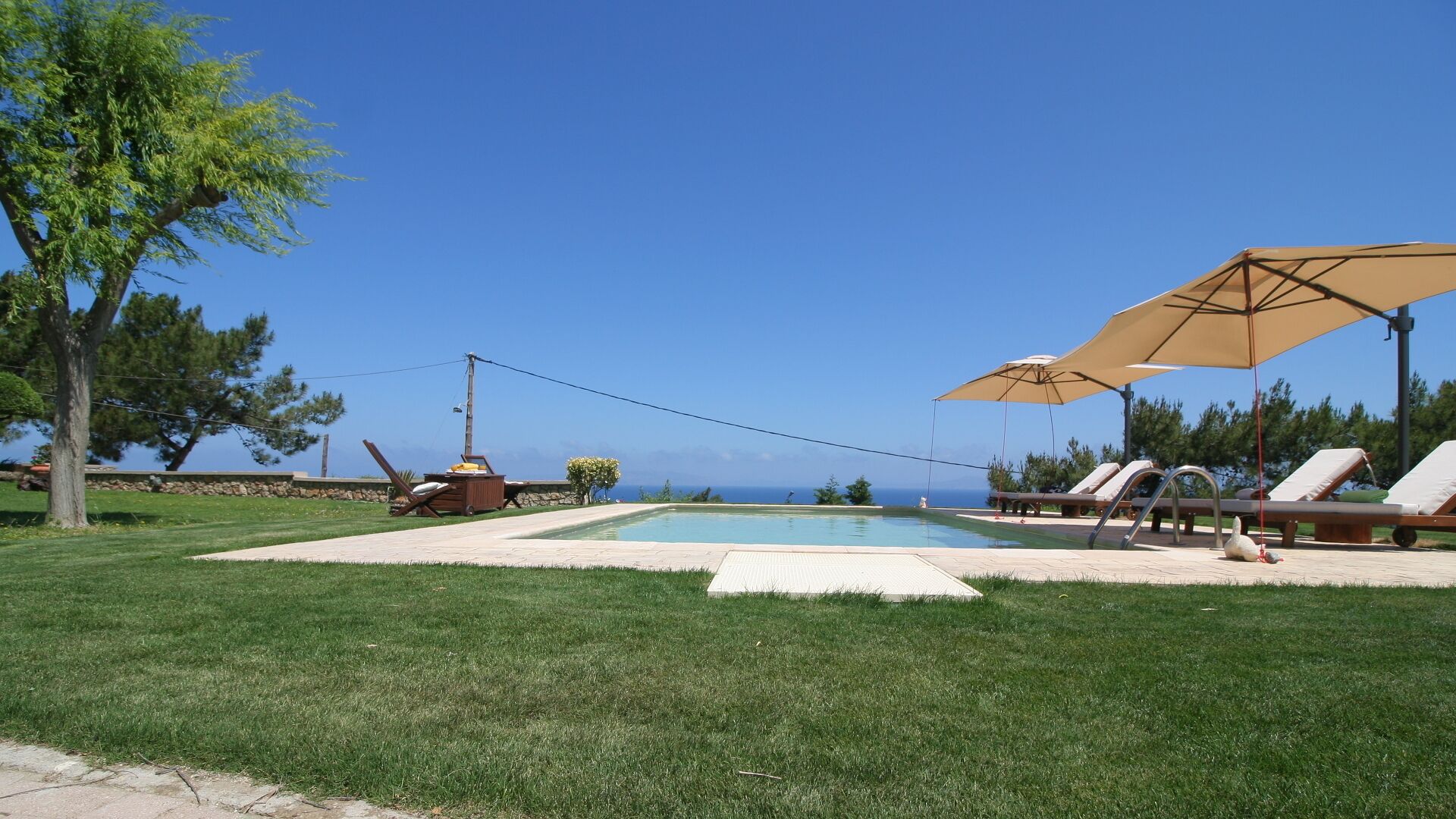 Una piscina con sillones de descanso y sombrillas, rodeada de césped cuidado y árboles.