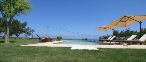 Una piscina con sillones de descanso y sombrillas, rodeada de césped cuidado y árboles.