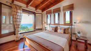 A bedroom with a wooden ceiling, a large bed with multiple pillows, a nightstand, a lamp, and a view of the ocean through a window. - Four-Bedroom Private Family Villa with Pool, 400m from the Beach. Villa Russelia (Kamiros skala)