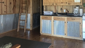 A rustic kitchen with wooden cabinets, a ladder, a coffee maker, and a small sink. - Cozy Cabin on the Creek with 2 private sandbars (Perkinston)