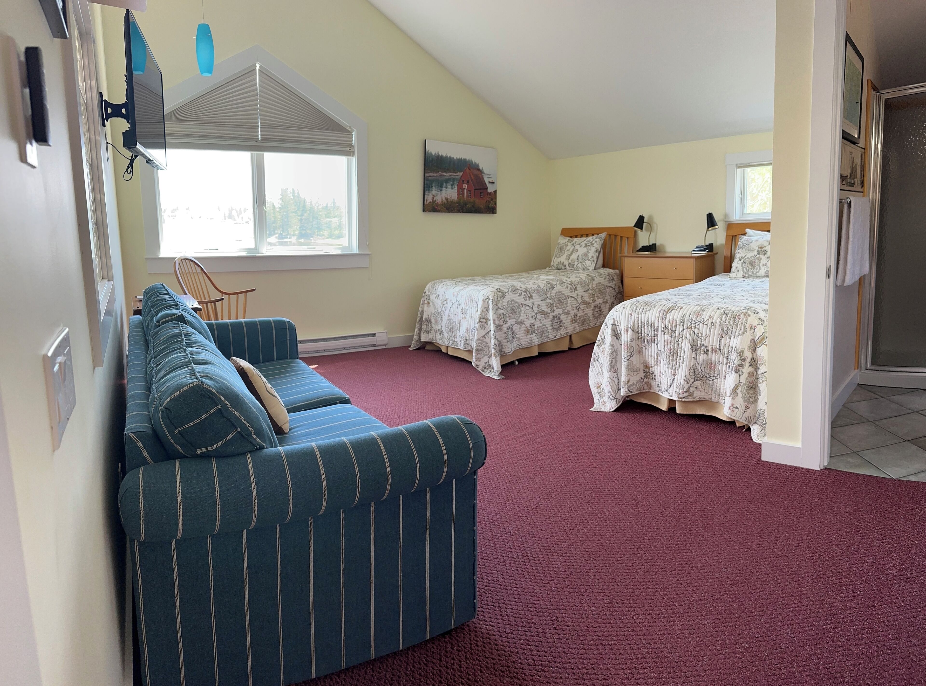 A bedroom with a sloped ceiling, two single beds, a blue striped sofa, a painting on the wall, and a window with blinds.