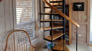 Una escalera de caracol con escalones de madera y barandillas de metal, una silla de madera y una estantería frente a un muro con paneles de madera.
