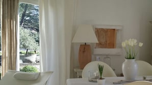 Una habitación con una mesa de comedor dispuesta para dos personas, una lámpara, un jarrón con flores blancas y vistas al exterior con vegetación.