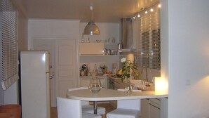 Una cocina compacta con encimera blanca, dos sillas blancas, una pequeña mesa de comedor, un refrigerador, un lavamanos y una lámpara colgante.
