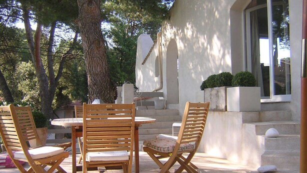 Un área de estar al aire libre con una mesa redonda y cuatro sillas de madera, rodeada de árboles y un edificio de fachada blanca con ventanas arqueadas.