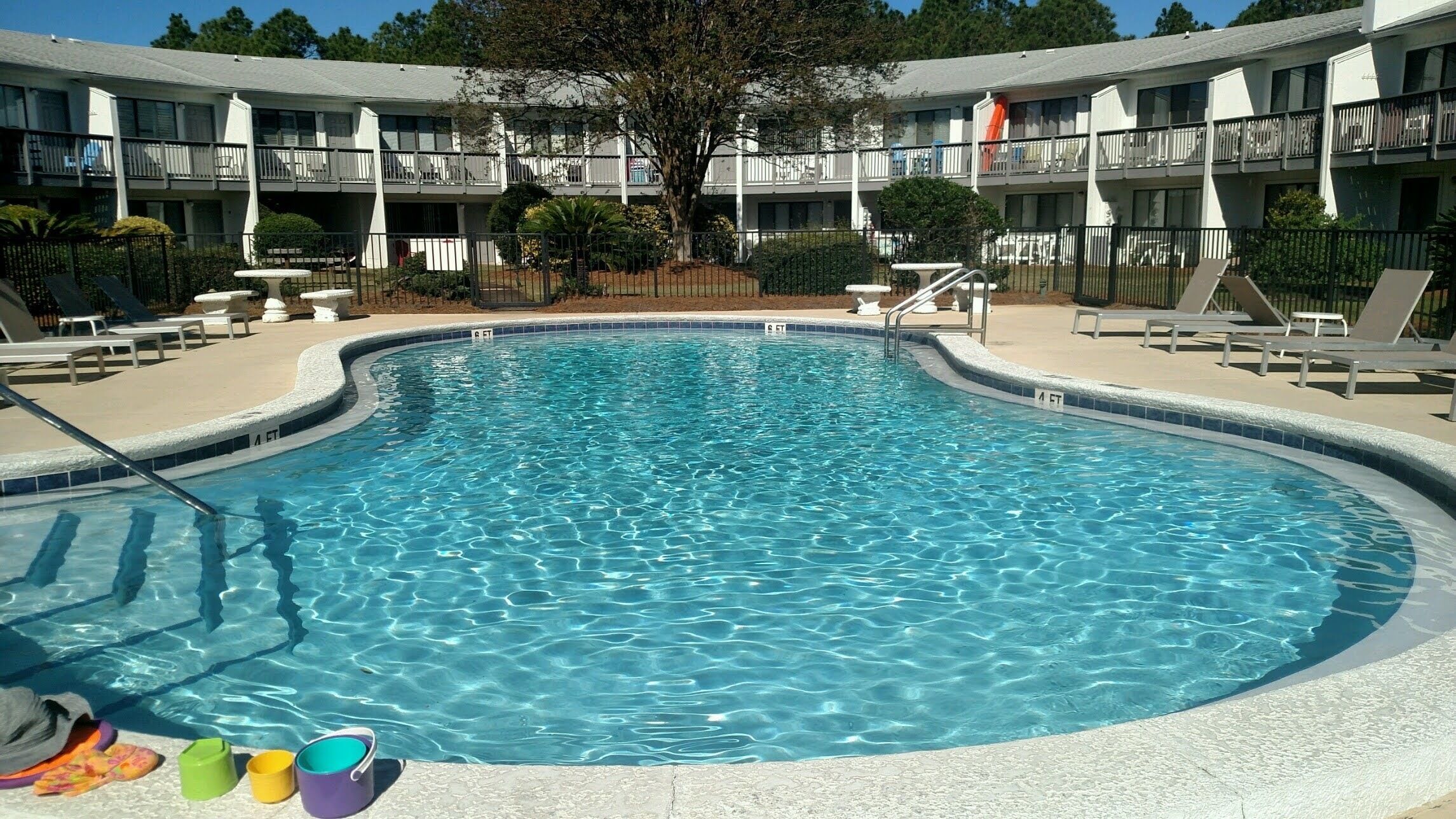 A swimming pool with a curved edge, surrounded by lounge chairs and a building with balconies in the background.