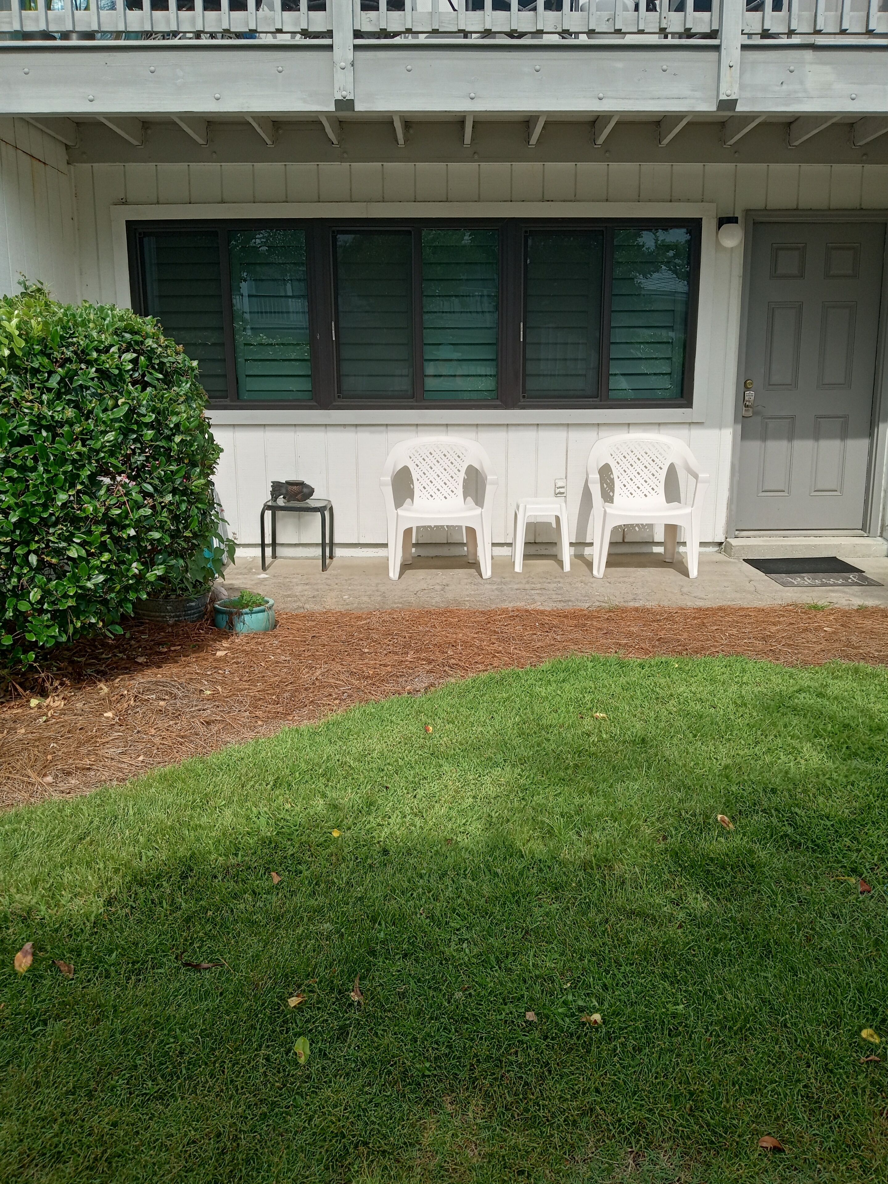 A house with a front porch, two white plastic chairs, a small table, and a green bush.